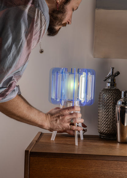A Mini Poppy lamp in Arctic Blue sits on a modern bedside table, glowing softly. Its sculptural shape and translucent acrylic surface add a calming, ambient light to the minimalist bedroom. Perfect for small space lighting and contemporary interior styling.

